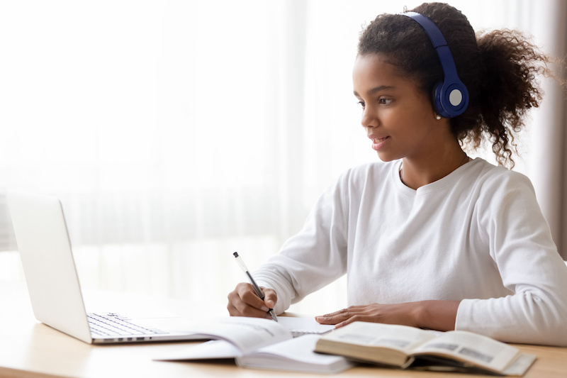 African American girl with headphones on is looking at her laptop and writing into a notebook, several books are lying open next to her